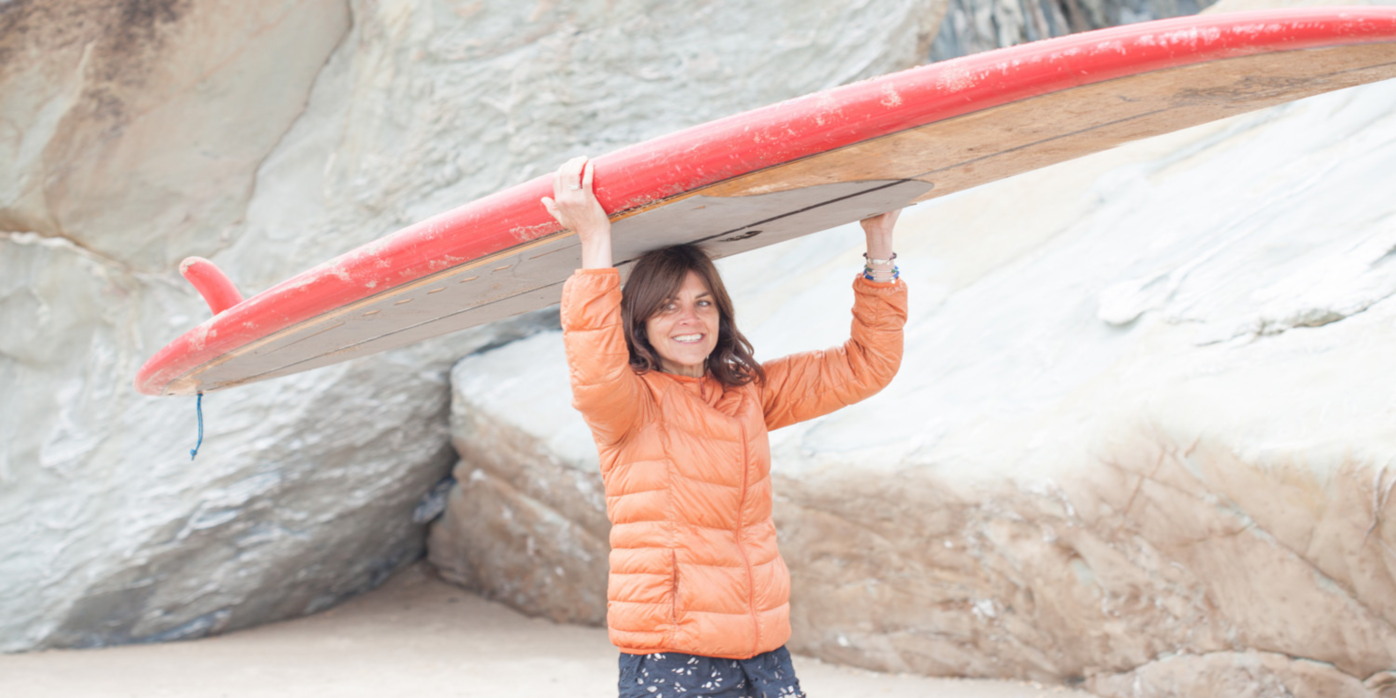 Helen Butler carrying a surfboard on the beach, reflecting the coastal inspiration behind Helen Butler Designs handmade silver jewellery