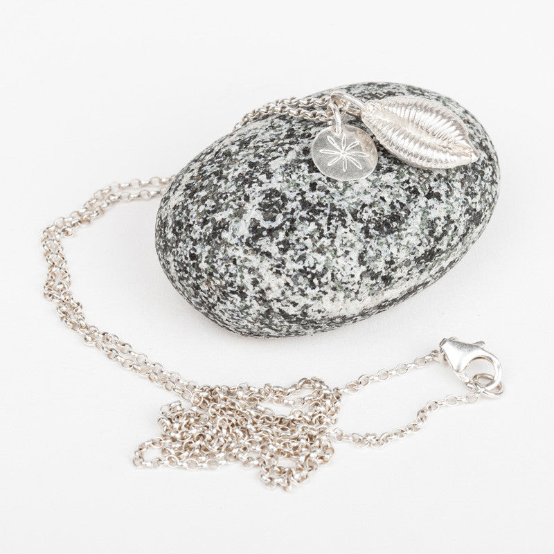 A sterling silver cowrie shell long pendant necklace with a fine silver chain laid out next to a rock.